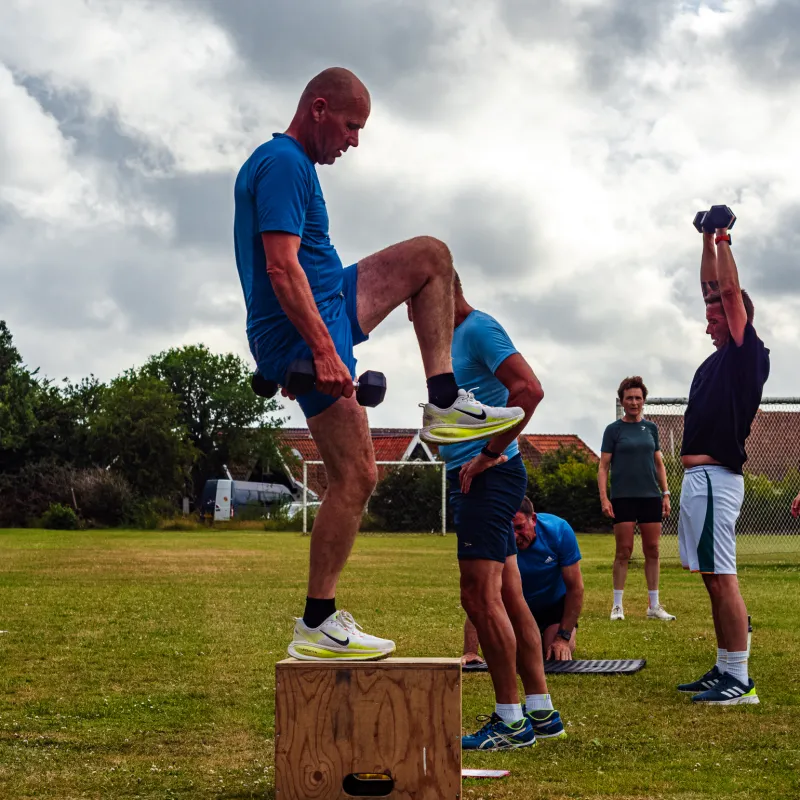 Deelnemers trainen buiten met box jumps en gewichten tijdens een groepsles van FitCoach Wilma op het grasveld.